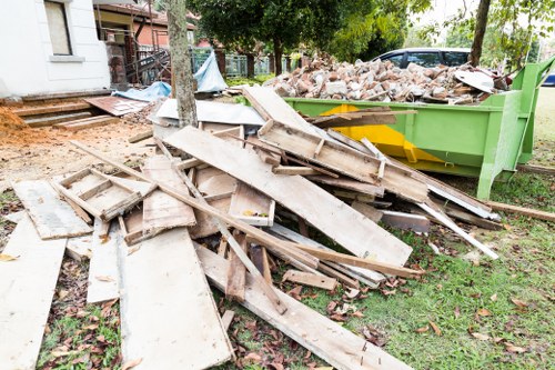 A pile of broken and discarded wooden door panels and planks, featuring weathered and chipped surfaces with pale, natural wood tones, resting on a grassy area near a green skip filled with brick rubble. The debris is partially overlapping and appears to have been recently removed from a residential property, with some boards leaning against a tree and scattered across the lawn. In the background, there are residential buildings with white exteriors and brown fencing, along with lush green trees and shrubs, indicating a suburban setting. The scene is illuminated by natural daylight, emphasizing the textures and colors of the waste materials. This image reflects typical rubbish removal activities handled by Hendon House Clearance in the local area, focusing on the disposal of construction debris and household waste.